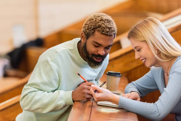 Positive interracial students looking at notebook near coffee to go in university — Stock Photo