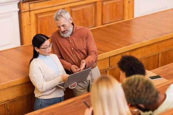 Maturo insegnante indicando laptop vicino asiatico studente in università — Foto stock