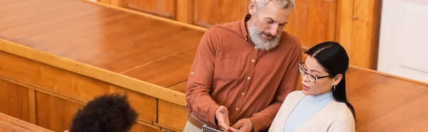 Middle aged teacher pointing at laptop near asian student in eyeglasses in university, banner — Stock Photo