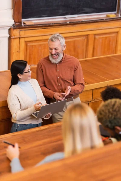 Sorridente insegnante che punta al computer portatile vicino studente asiatico e persone offuscate in università — Foto stock