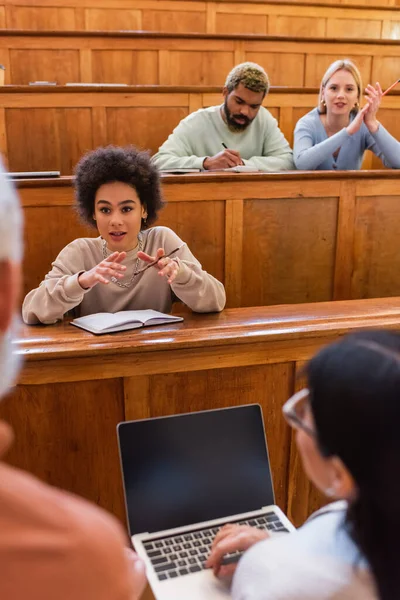 Estudiante afroamericano hablando con profesor borroso y amigo con portátil en la universidad - foto de stock