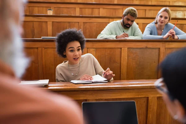 Joven estudiante afroamericano con cuaderno hablando con el profesor y amigo cerca de la computadora portátil en la universidad - foto de stock