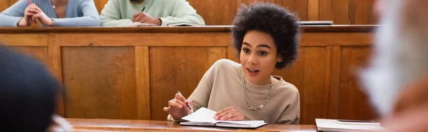 Étudiant afro-américain avec cahier parlant à un professeur flou à l'université, bannière — Photo de stock