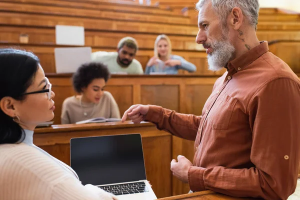 Mature teacher pointing at laptop with blank screen near asian student in university — Stock Photo