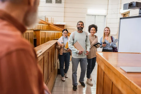 Cheerful multicultural students with notebooks and devices standing near blurred teacher in university — Stock Photo