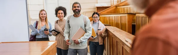 Estudiantes interracial positivos con cuadernos y dispositivos mirando al profesor borroso en la universidad, pancarta - foto de stock