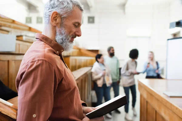 Mature teacher holding notebook near blurred interracial students in university — Stock Photo
