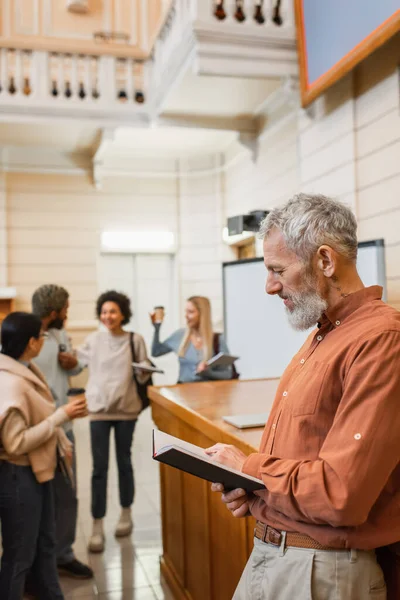 Profesor de mediana edad sosteniendo cuaderno cerca de estudiantes interracial en la universidad - foto de stock