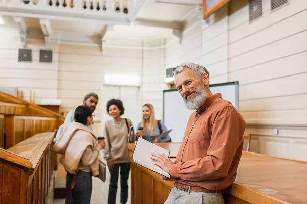 Profesor sonriente sosteniendo un cuaderno y mirando a la cámara cerca de estudiantes multiétnicos borrosos en la universidad - foto de stock