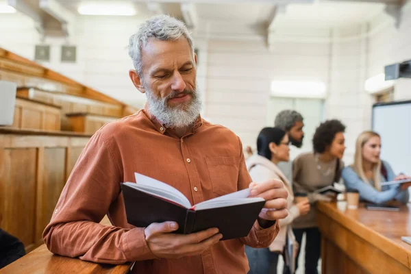 Lehrer mittleren Alters hält Notizbuch in der Nähe verschwommener multiethnischer Studenten an der Universität — Stockfoto