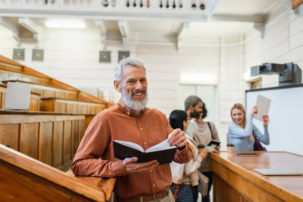 Positivo insegnante maturo in possesso di quaderno vicino agli studenti offuscati in auditorium universitario — Foto stock