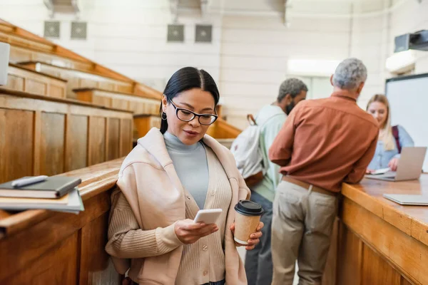 Étudiant asiatique utilisant un smartphone et tenant du café pour aller près des amis flous et professeur à l'université — Photo de stock