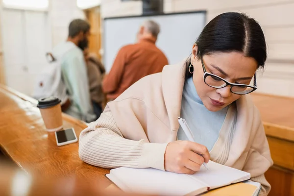Asian student writing on notebook near smartphone and coffee to go in university — Stock Photo