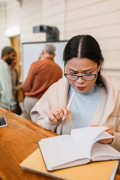 Pensive asiatique étudiant à la recherche à notebook près smartphone dans auditorium universitaire — Photo de stock