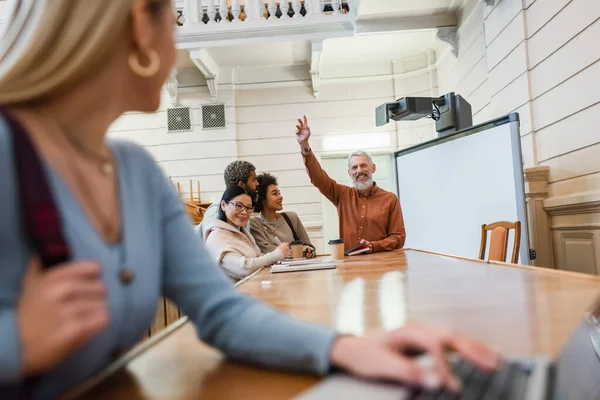 Smiling teacher waving hand at blurred student with laptop in university — Stock Photo