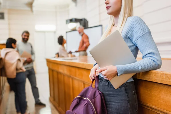 Estudiante rubia sosteniendo mochila y portátil cerca de amigos borrosos en la universidad - foto de stock