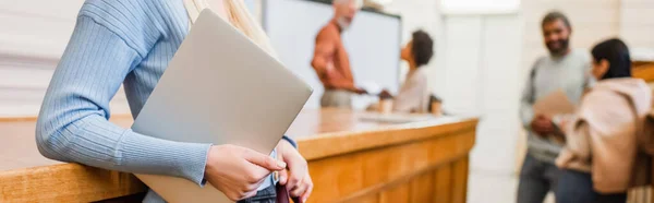 Student holding laptop near blurred friends in university, banner — Stock Photo