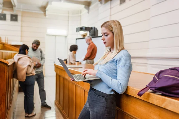 Étudiant blonde utilisant un ordinateur portable près du sac à dos et des amis interraciaux à l'université — Photo de stock