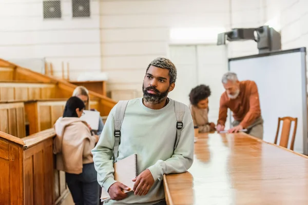 African american student holding notebook near blurred friends and teacher in university — Stock Photo