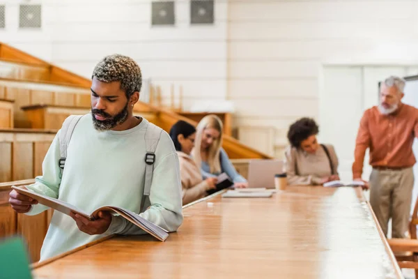 Estudante afro-americano segurando notebook perto de amigos turvos e professor no auditório universitário — Fotografia de Stock