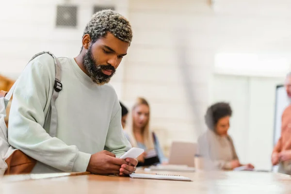 Studente afroamericano con zaino utilizzando smartphone vicino notebook all'università — Foto stock