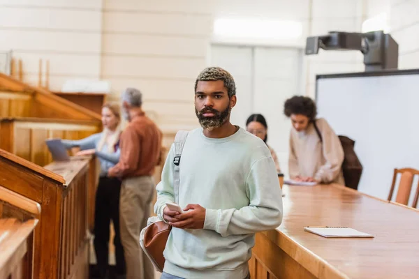 African american student with backpack and smartphone looking at camera in university — Stock Photo