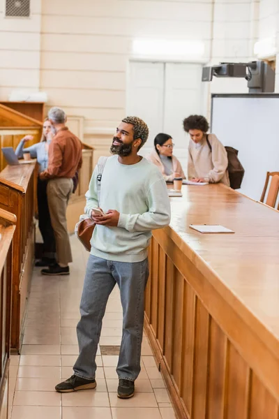 Sourire étudiant afro-américain avec sac à dos tenant smartphone à l'université — Photo de stock