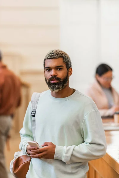 Jeune étudiant afro-américain avec sac à dos et téléphone portable regardant la caméra à l'université — Photo de stock