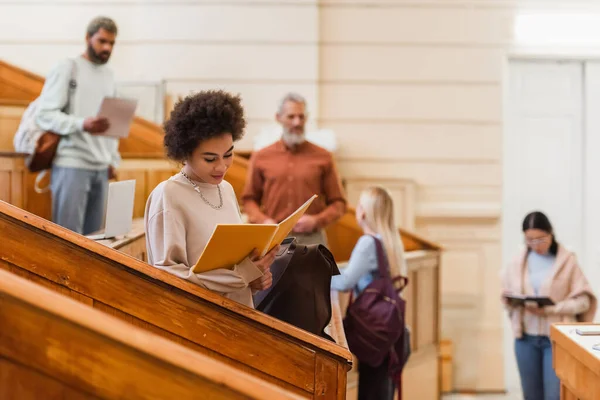 Afroamerikanische Studentin schaut in der Nähe verschwommener Freunde und Lehrer auf Notizbuch — Stockfoto