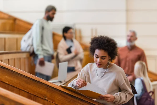 African american student looking at notebook near blurred friends and teacher in university — Stock Photo