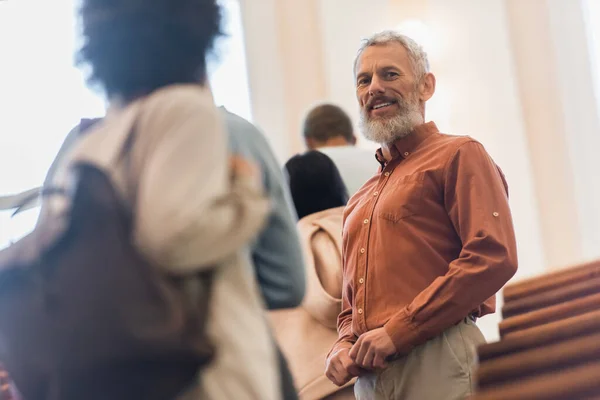 Vista de ángulo bajo del profesor sonriente mirando a la cámara cerca de estudiantes interracial borrosas en la universidad - foto de stock