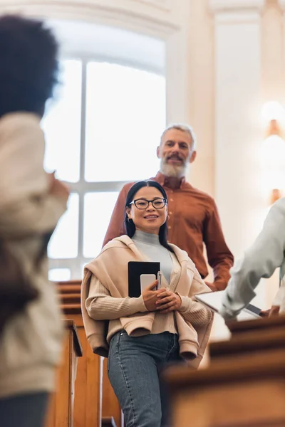 Low angle view of smiling asian student with smartphone and notebook looking at camera near blurred friends and teacher in university — Stock Photo