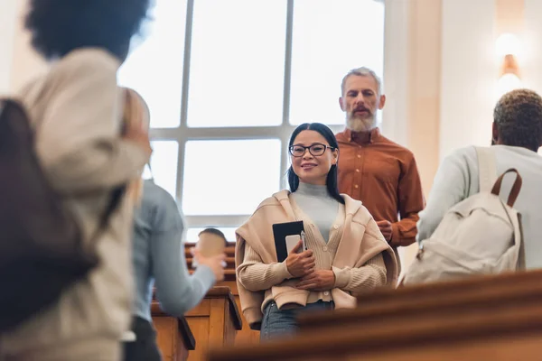 Позитивний азіатський студент тримає блокнот і смартфон біля міжрасових друзів і вчителя в університеті — стокове фото
