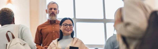 Feliz asiático estudiante con notebook mirando cámara cerca borrosa profesor y interracial amigos en la universidad, banner - foto de stock