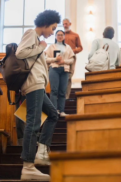 African american student with backpack and notebook standing on stairs in university — Stock Photo