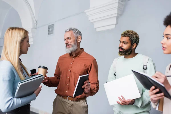 Profesora madura sosteniendo cuaderno cerca de estudiantes interracial con dispositivos y café para ir en corredor universitario - foto de stock