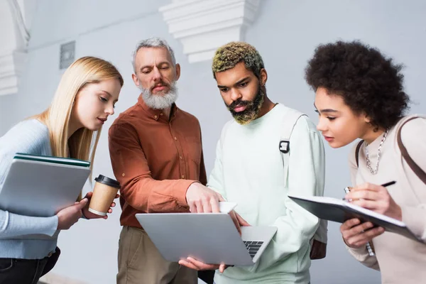 Middle aged teacher with cellphone standing near multiethnic students with laptops in university corridor — Stock Photo