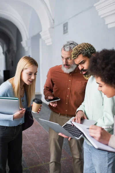Teacher holding smartphone and talking to multiethnic students with laptops and notebooks in university — Stock Photo