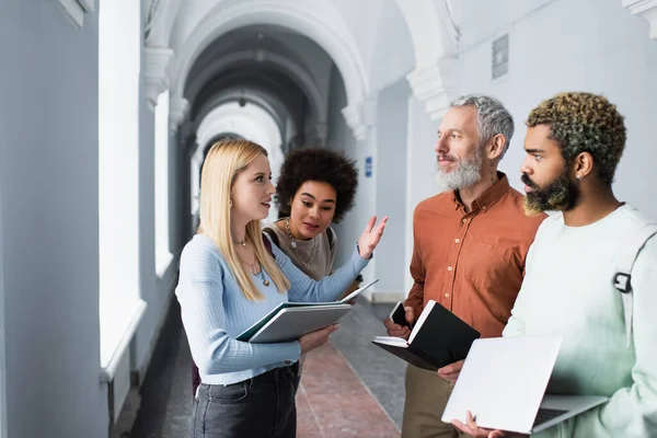 Happy student with laptop talking to teacher and african american friends in university corridor — Stock Photo