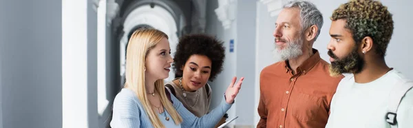 Smiling student pointing with hand near teacher and african american friends in university corridor, banner — Stock Photo