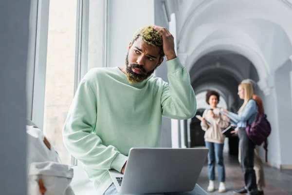 Perturbado estudiante afroamericano usando portátil cerca de ventana en corredor universitario - foto de stock