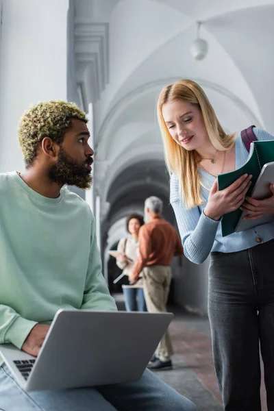 Lächelnder Student hält Notizbuch in der Nähe afrikanisch-amerikanischer Studentin mit Laptop auf Universitätsflur — Stockfoto