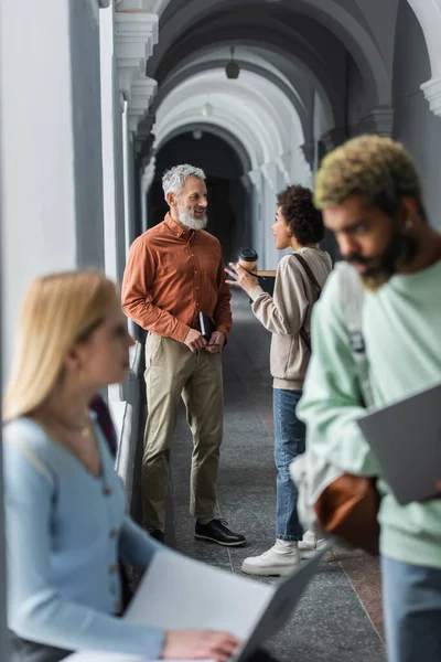 Cheerful teacher holding notebook near african american student with coffee to go in university corridor — Stock Photo