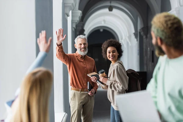 Professeur souriant agitant la main aux étudiants interracial dans le couloir de l'université — Photo de stock