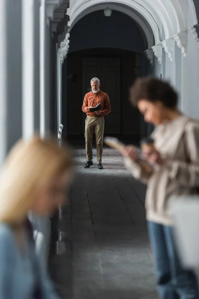 Professeur mature regardant un cahier près des étudiants interracial flous dans le couloir de l'université — Photo de stock