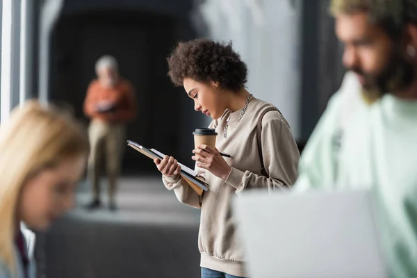 Giovane studente afroamericano in possesso di caffè per andare e quaderni vicino amici all'università — Foto stock