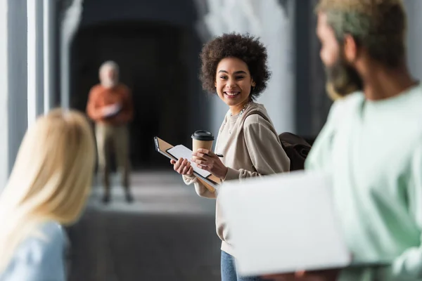 Sorridente studente africano americano in possesso di caffè per andare e quaderni vicino amici offuscati in università — Foto stock