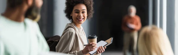 Étudiant afro-américain joyeux tenant du café à emporter et des cahiers près d'amis flous à l'université, bannière — Photo de stock