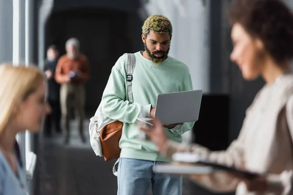 Studente afroamericano con zaino utilizzando laptop vicino ad amici sfocati nel corridoio universitario — Foto stock