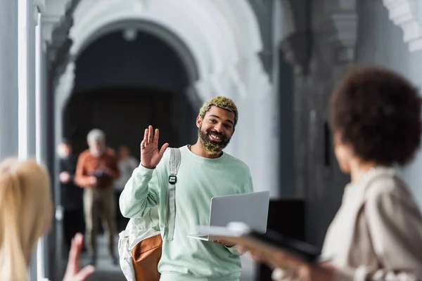African american student with laptop waving hand at blurred friends in university corridor — Stock Photo